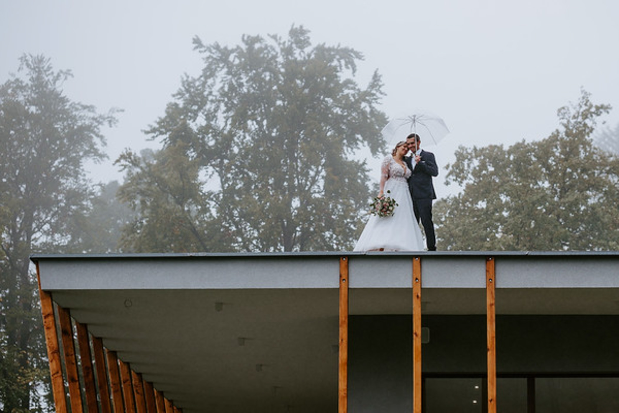 Novomanžele na střeše slavnostního sálu Chaty Dukla — romantický snímek s deštníkem a mlhavými horami